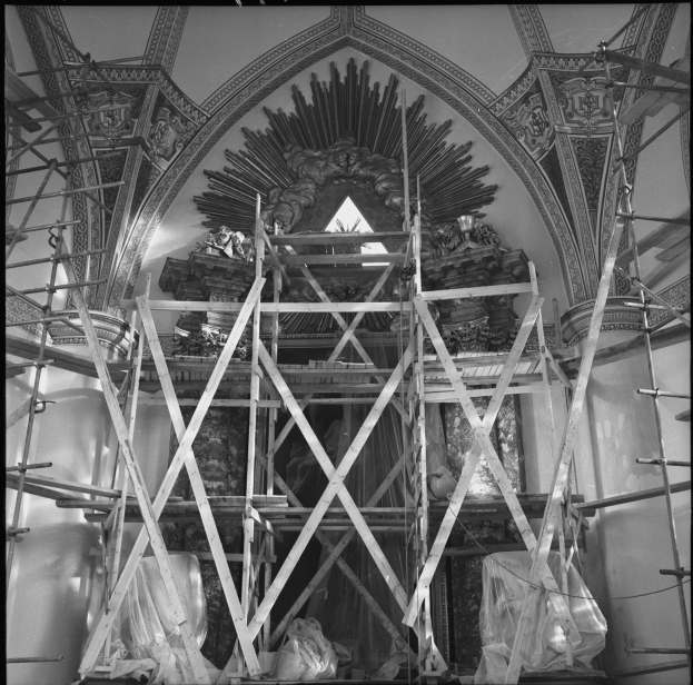 Schwarz-weiß-Foto einer Kirche mit Gerüst drumherum, das ein kunstvoll verziertes Gewölbe und sichtbare Renovierungsmaterialien an seiner Basis zeigt.