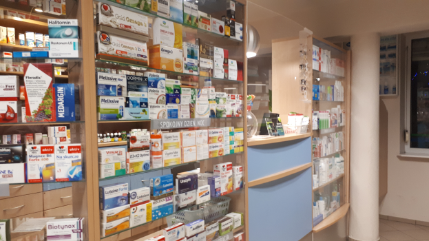 Eine Apotheken-Inneneinrichtung mit Regalen, die mit verschiedenen Medikamenten gefüllt sind, einer zentralen Säule und einem Glasfenster auf der rechten Seite.