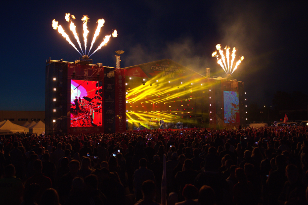 Eine große Menschenmenge auf einem Musikfestival schaut auf einer hell erleuchteten Bühne mit Bannern, umgeben von Zelten und Feuerwerk am dunklen Himmel.