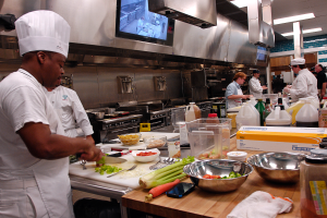 Eine Gruppe von Köchen in einer Küche, die auf einem Schneidbrett Lebensmittel zubereiten, mit einem Koch im Vordergrund in einer weißen Schürze und Mütze, der ein Messer hält und von verschiedenen Utensilien, Schüsseln, Gemüse, einem Handy, Flaschen und anderen Gegenständen umgeben ist, mit einem Fernseher und Deckenleuchten im Hintergrund.
