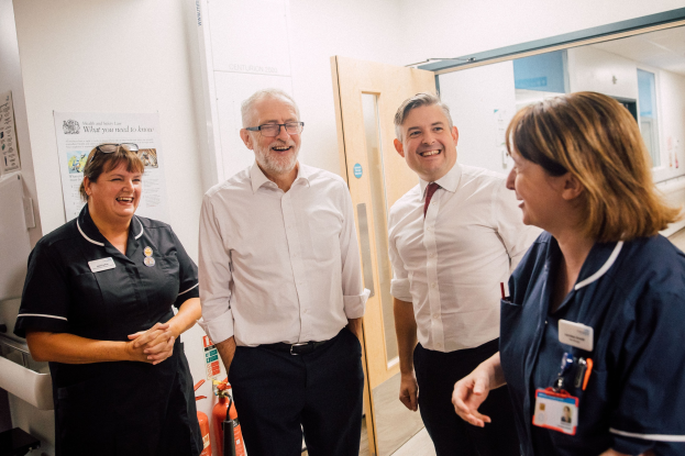 Vier Personen, die in einem Krankenhausflur mit Tür, Plakat, Feuerlöscher, Waschbecken, Wasserhahn und Spiegel im Hintergrund lächeln und sich unterhalten.