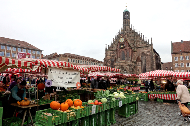 Ein belebter Markt in Nürnberg, Deutschland, mit farbenfrohen Obst- und Gemüsesorten, Menschen mit Taschen, Zelten und einem Uhrenturm im Hintergrund unter einem sichtbaren Himmel.