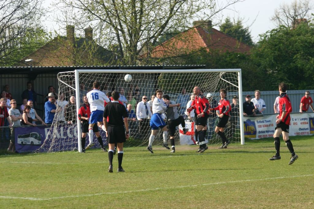 Spieler spielen F├╝├čball auf einem Feld mit einem Tor, w├Ąhrend Zuschauer dahinter stehen, mit B├Ąumen und H├Ąusern im Hintergrund.