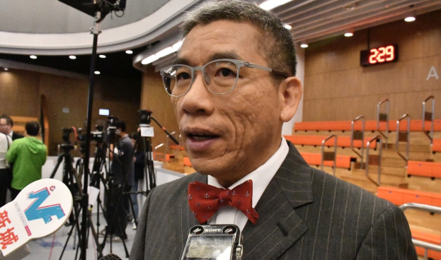 Ein Mann im Anzug mit Schleife spricht in ein Mikrofon, umgeben von Reportern und Kameras in einem beleuchteten Innenraum mit Bänken und einer Digitaluhr an der Wand.