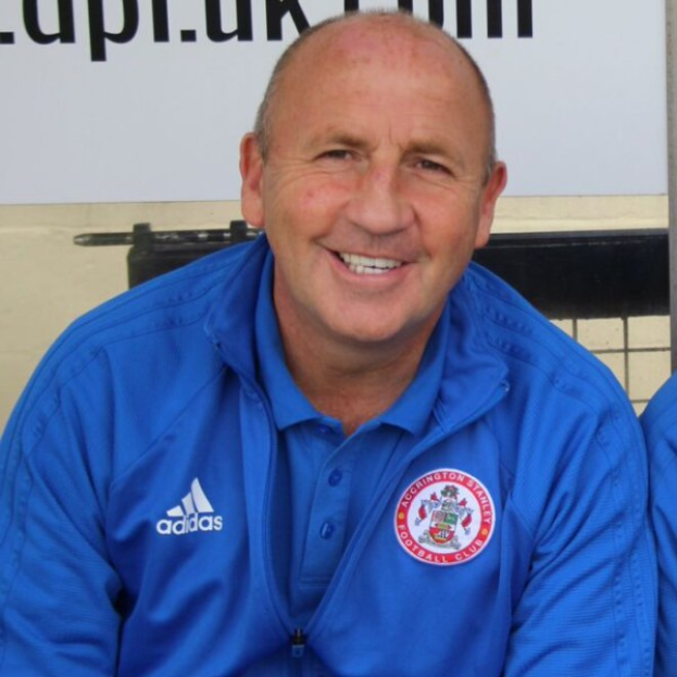 Zwei Männer in einem Raum, einer in einer blauen Jacke neben dem anderen sitzend, beide lachend, mit einer Wandtafel im Hintergrund.