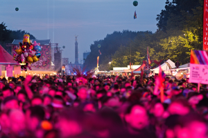 Große Menschenmenge auf einem nächtlichen Musikfestival mit farbigen Lichtern, Ballons, Zelten, Bannern, Fahnen, Bäumen, einem Turm und einem sternenklaren Himmel im Hintergrund.