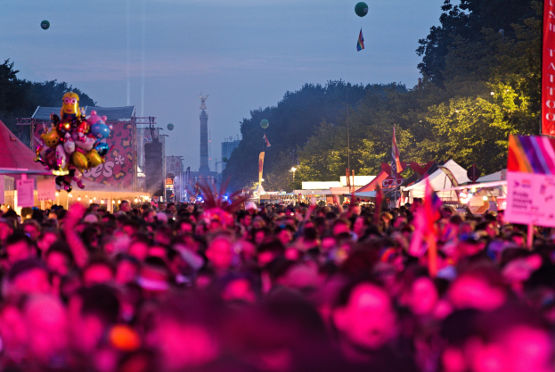 Große Menschenmenge auf einem nächtlichen Musikfestival mit farbigen Lichtern, Ballons, Zelten, Bannern, Fahnen, Bäumen, einem Turm und einem sternenklaren Himmel im Hintergrund.