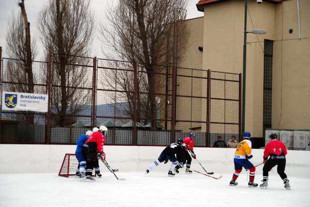 Menschen, die Eis-hockey auf einem Eisplatz mit Gebäuden, Bäumen, einer Straßenlaterne, einem Namensschild und Zäunen im Hintergrund und einem klaren Himmel spielen.