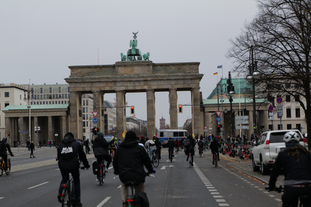 Eine Gruppe von Menschen, die auf Fahrr√§dern die Stra√Ÿe vor dem Brandenburger Tor in Berlin, Deutschland, entlangf√§hrt, mit B√§umen, Laternenm√§sten, Verkehrsampeln, Verkehrszeichen und Geb√§uden mit Fenstern im Hintergrund und dem Himmel in der Ferne.