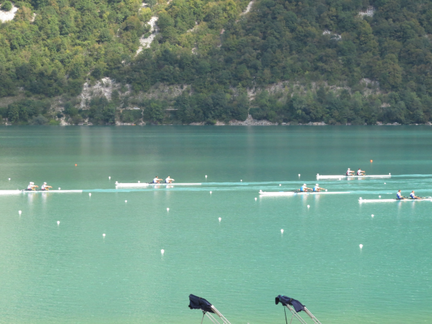 Gruppe von Menschen beim Ruderwettbewerb auf einem Gewässer mit Bäumen und einem Hügel im Hintergrund, Kleidung auf Ständen am Boden.