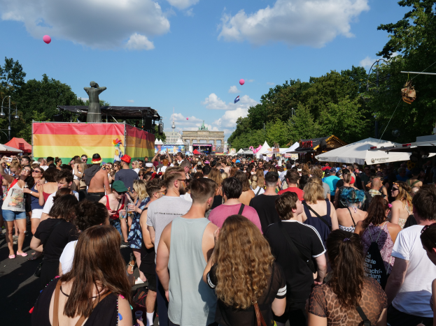 Eine große Menschenmenge, die eine Straße mit Zelten, Bäumen, Pfählen, Lichtern, einer Statue und Gebäuden im Hintergrund entlanggeht, unter einem bewölkten Himmel mit Ballons, beim Christopher Street Day in Berlin.