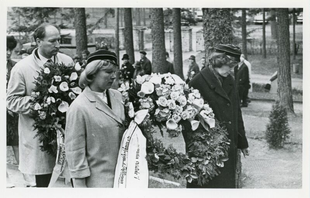 Eine Gruppe von Menschen steht nebeneinander, jeder hält einen Blumenstrauß in der Hand, auf einem Schwarz-Weiß-Bild mit Bäumen und Gebäuden im Hintergrund, wahrscheinlich bei einer Holocaust-Gedenkfeier.
