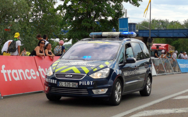Ein Polizeiwagen fährt die Straße entlang neben einer Menge, mit einigen Leuten, die Mötzen tragen und Taschen haben, einem Banner links, Gelöndern mit Bannern dahinter und einem Hintergrund mit Bäumen, einer Brücke, einer Fahne auf einem Mast und einem bewölktem Himmel.