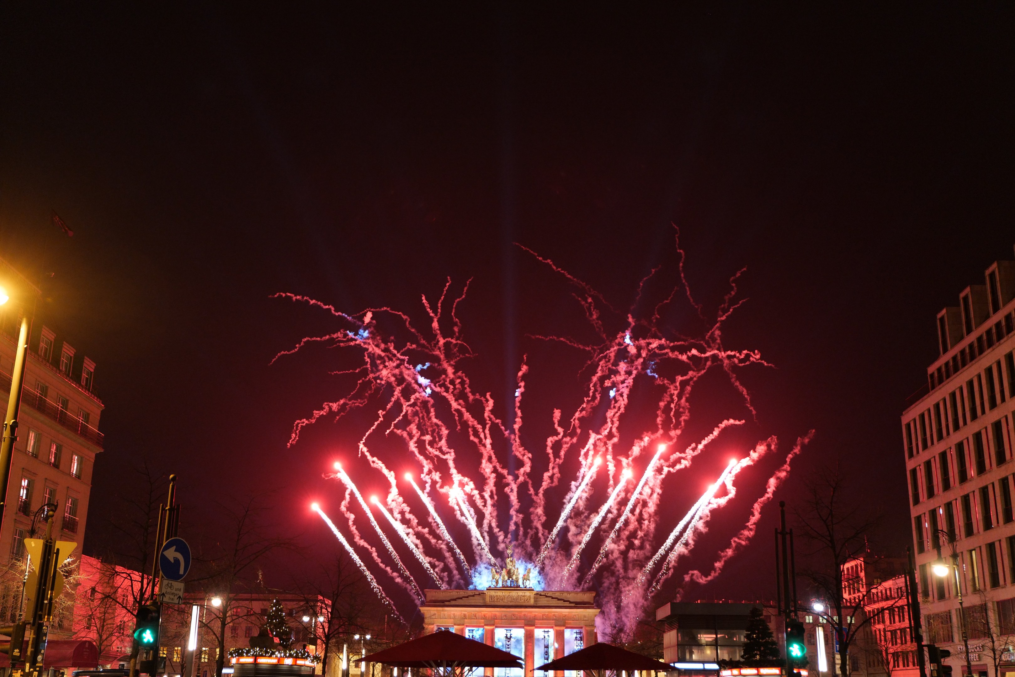 Eine belebte Stadtstraße am Neujahr in Berlin, voller Menschen, Fahrzeuge und Gebäude, beleuchtet von Lichtern und Feuerwerken am Himmel.