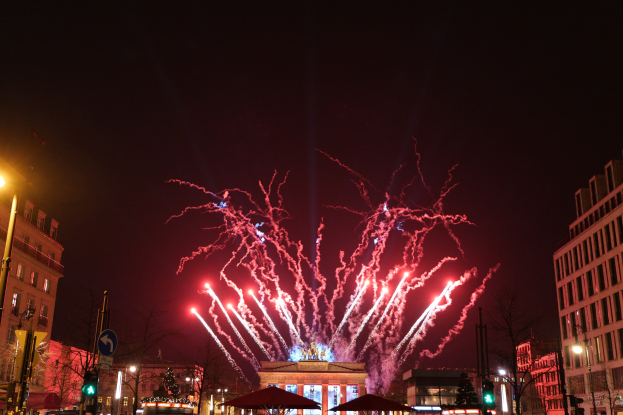 Eine belebte Stadtstraße am Neujahr in Berlin, voller Menschen, Fahrzeuge und Gebäude, beleuchtet von Lichtern und Feuerwerken am Himmel.