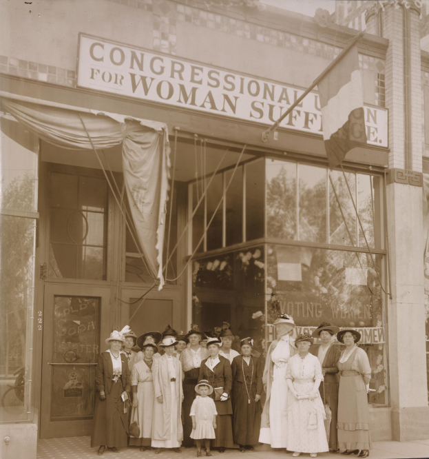 Eine Gruppe von Frauen in Kleidern, einige mit Hüten, steht vor einem Gebäude mit einer "Congressional for Woman Suffrage"-Aufschrift, Glaswänden, einer Tür und einer Texttafel.