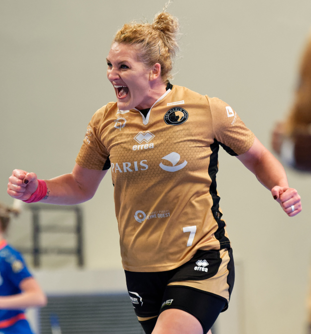 Eine Frau in braunem T-Shirt und schwarzen Shorts rennt auf einem Futsal-Platz und feiert mit triumphaler Miene, während zwei andere Frauen im Hintergrund unscharf sind.