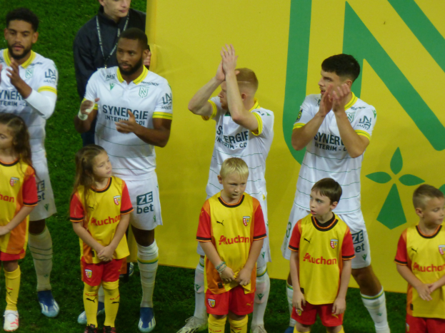 Eine Gruppe junger Menschen in Fußballtrikots steht auf einem Fußballfeld und klatscht feiernd mit einer 'Ligue 1'-Plakette im Hintergrund.