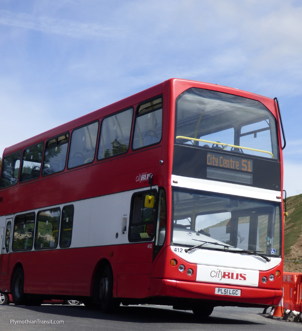 Ein roter Doppeldecker-Stadtbus fährt mit einem Verkehrskegel in der Nähe und einem Hügel im Hintergrund.