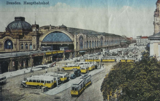 Ein sepiafarbenes Postkartenbild des Dresdner Hauptbahnhofes in Deutschland, das eine belebte Straßen Szene mit Fahrzeugen, Fußgängern, Bäumen, Gebäuden und einem bewölkten Himmel zeigt.