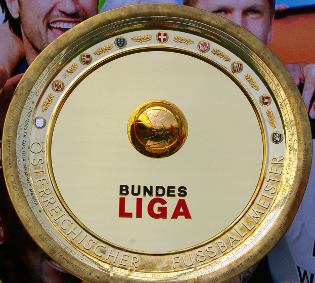 Ein Mann hält einen goldenen Schild mit der Aufschrift "Bundesliga" hoch, im Hintergrund eine Fahne mit drei Personen.