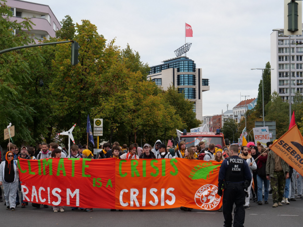 Eine Gruppe von Menschen marschiert eine von Bäumen gesäumte Straße entlang und hält ein Banner mit der Aufschrift "Klimakrise ist eine Krise", mit parkenden Fahrzeugen, Gebäuden und einem klaren blauen Himmel im Hintergrund.