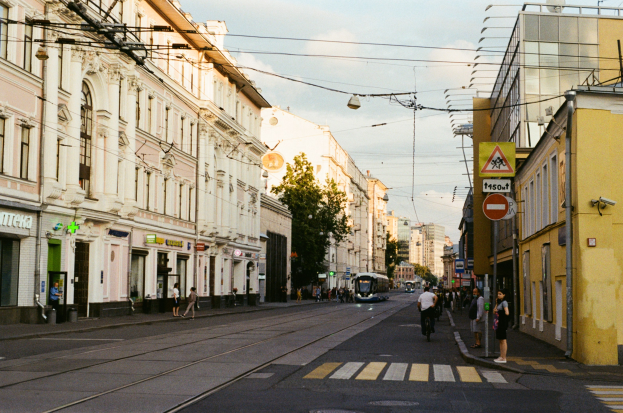 Stadtstraße mit Fußgängern und Radfahrern, umgeben von Gebäuden, Schildern, Bäumen und Strommasten mit Drähten unter einem bewölkten Himmel.