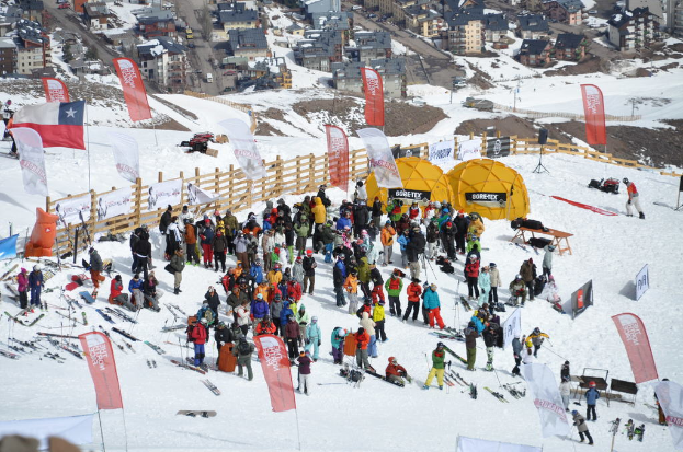 Menschen stehen und sitzen auf Schnee, einige Schlittschuhlaufen, mit einem hölzernen Zaun, der Tafeln mit Text und Bildern trägt, Flaggen, Zelten und verschiedenen Gegenständen im Hintergrund.