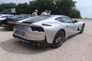 Roter Ferrari 812 Superfast auf dem Goodwood Festival of Speed umgeben von Menschen mit Mützen, Bäumen, Texttafeln und einem bewölkten Himmel im Hintergrund.