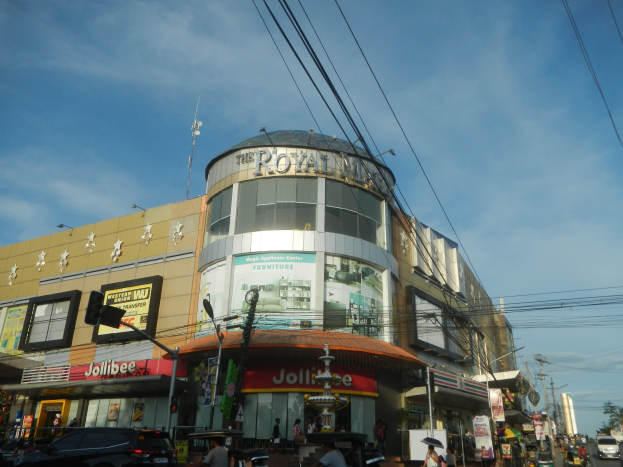 Eine belebte Stadtstraße mit fahrenden Autos, Fußgängern auf dem Gehweg und dem Royal Mall Einkaufszentrum im Hintergrund, umgeben von Bäumen, Strommasten und Drähten unter dem Himmel.