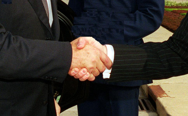 Zwei Männer im Business-Outfit schütteln sich die Hand vor einem Gebäude, umgeben von grünem Gras und Pflanzen, was eine formelle Geschäftsbesprechung suggeriert.