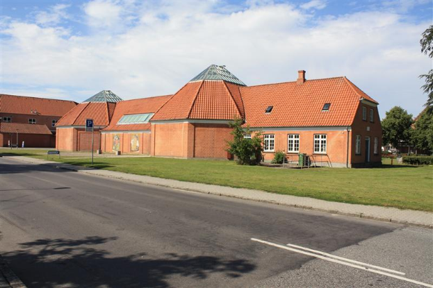 Ein großes Backsteingebäude mit rotem Dach, umgeben von Grünflächen und Schildern, das an der Seite einer Straße steht, unter einem bewölkten Himmel, das die neue Schule in den Niederlanden darstellt.