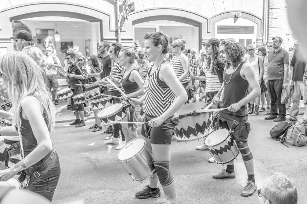 Eine Gruppe von Menschen, die draußen vor einer geschlossenen Glastür mit Licht im Hintergrund Musikinstrumente spielen.