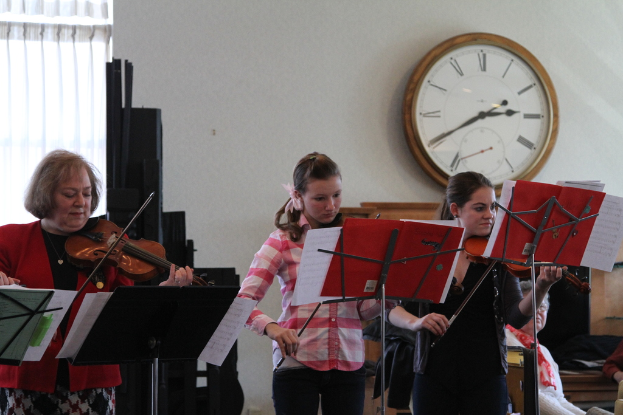 Drei Frauen auf der Bühne mit Geigen stehend und auf Musikständern spielend, während eine Gruppe sitzender Frauen im Vordergrund zuhört; eine Uhr hängt an der weißen Wand im Hintergrund, mit einem weißen Vorhang in der linken Ecke.