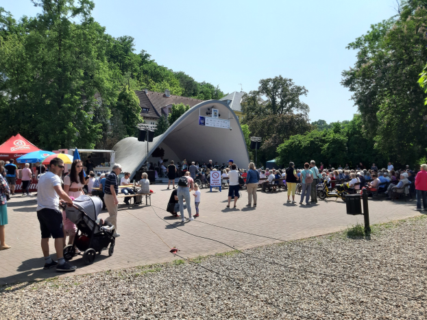 Eine Menschenmenge versammelt sich vor einem Gebäude bei der Eröffnung eines Festivals, wobei einige auf Bänken sitzen und andere stehen, einschließlich einer Person mit einem Kinderwagen im Vordergrund. Bäume, Gebäude und ein klarer blauer Himmel bilden den Hintergrund, während Zelte die linke Seite säumen und ein Müllcontainer auf der rechten Seite zu sehen ist.
