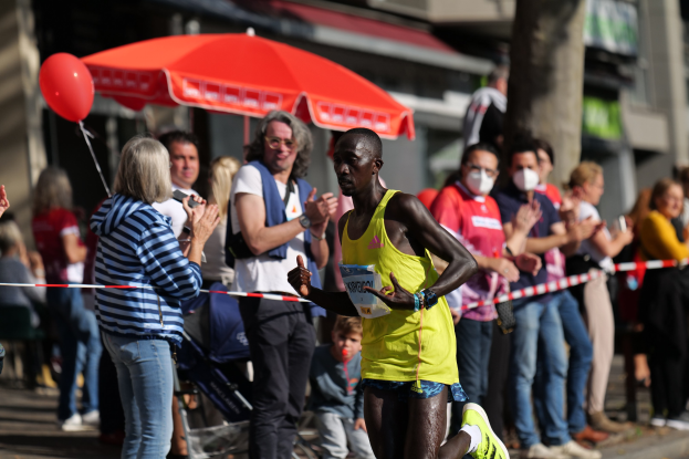 Ein Mann in einem gelben T-Shirt und Laufschuhen sprintet durch einen Marathon, während ihn eine Menge anfeuert, mit einem Schirm und einem Kinderwagen im Hintergrund.
