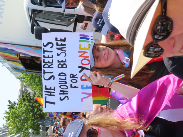 Eine Gruppe von Menschen hält ein "Die Straßen sollten sicher sein"-Schild vor einem Lastwagen, einige tragen Mützen und Schutzbrillen, andere Schilder, vor einem Hintergrund aus Bäumen, Gebäuden und einem klaren blauen Himmel.