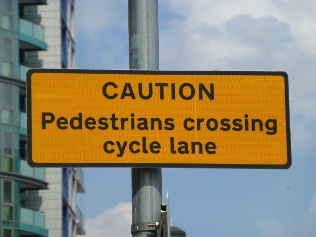 Gelbes Warnschild an einem Pfosten mit der Aufschrift "Fußgänger überqueren Fahrradspur" vor einem Gebäude vor einem klaren blauen Himmel.