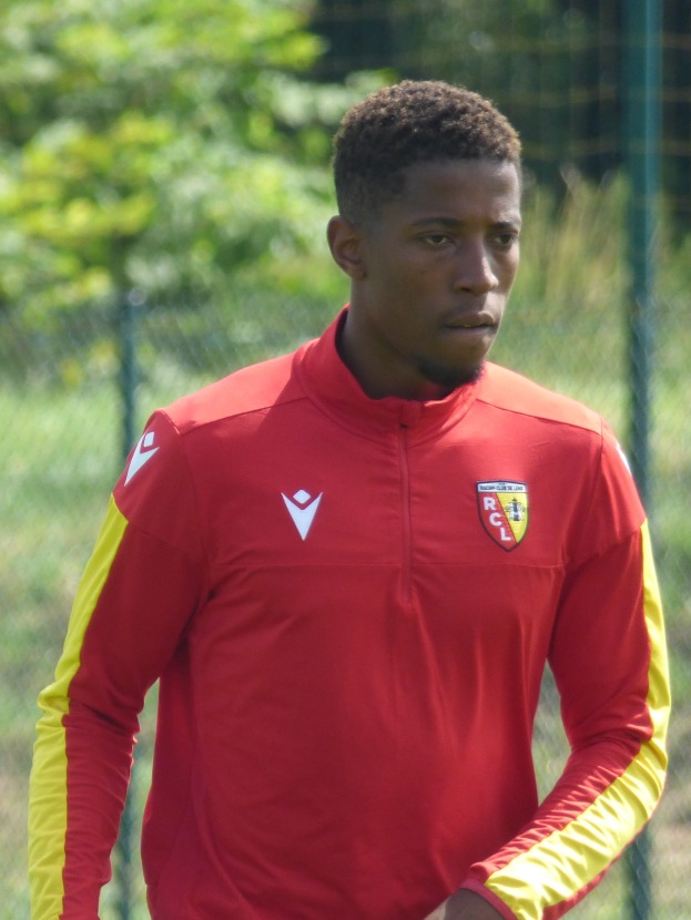 Ein junger Mann in einem roten und gelben Trikot steht auf einem Fußballfeld, umgeben von einem Zaun und Bäumen, mit einem entschlossenen Gesichtsausdruck, wahrscheinlich ein Flügelstürmer.