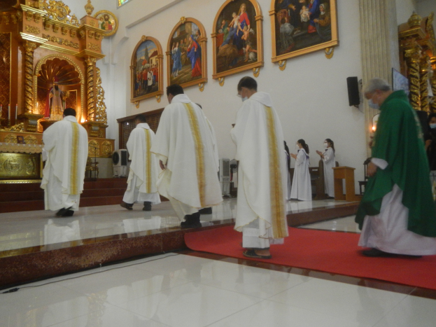 Eine Gruppe von Priestern geht einen roten Teppich in einer Kirche entlang, während Menschen auf dem Boden vor ihnen und auf der rechten Seite stehen und Fotoalben an der Wand im Hintergrund hängen.