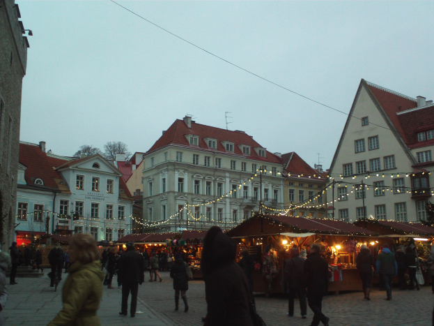 Ein belebter Weihnachtsmarkt in Tallinn, Estland mit Menschen, beleuchteten Ständen, Gebäuden, Bäumen und einem klaren blauen Himmel, mit Text am unteren Rand.