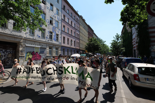 Eine Gruppe von Menschen marschiert auf einer Straße mit einem Banner, rechts parkende Autos, links ein Fahrradfahrer und im Hintergrund Gebäude, Bäume und ein Schild unter einem sichtbaren Himmel.