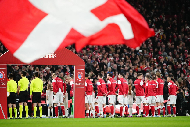 Eine Gruppe von Menschen auf einem Fußballfeld mit einer roten und weißen Flagge im Vordergrund, einem Bogen mit dem Text "Bayern Munich vs Bayern Munich Betting Tips & Preview" im Hintergrund und einer großen Menschenmenge im Stadion.
