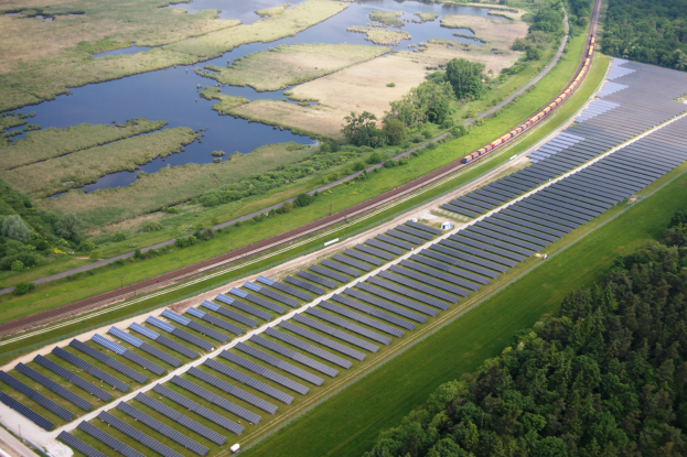Eine Luftaufnahme eines Solarparks mit zahlreichen Solarpanelen in einem Feld, umgeben von Bäumen, Gras, Wasser und einer nahen Bahnlinie.