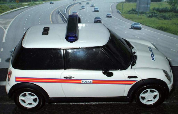 Weißes Polizeifahrzeug mit "Polizei"-Text darauf, gefolgt von mehreren Fahrzeugen auf einer Straße mit Gras und Bäumen außerhalb der Stadt.