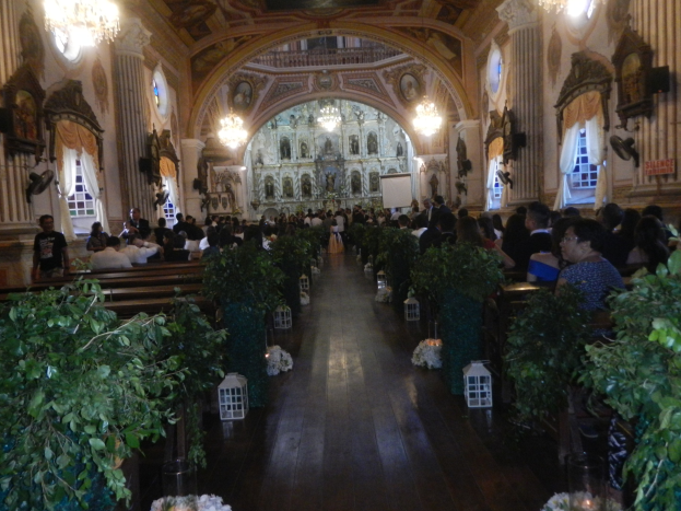 Eine Trauung in einer Kirche mit Gästen auf Bänken und im Gang sitzend und stehend, dekoriert mit Zimmerpflanzen, Skulpturen, Vorhängen und Kronleuchtern.