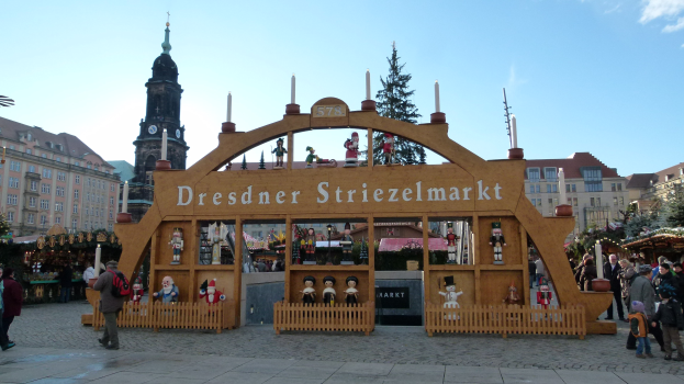Ein geschäftiger Weihnachtsmarkt in Dresden, Deutschland mit Menschen, die spazieren gehen, einem Torbogen, Bäumen, Gebäuden und einem bewölkten Himmel.
