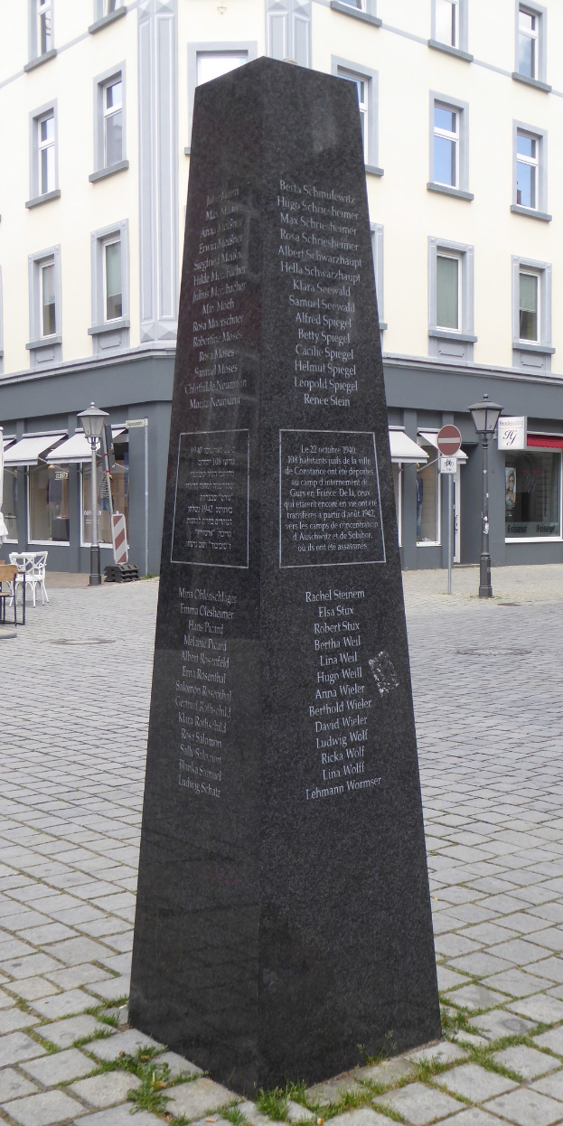 Ein Denkmal mit Inschrift steht in einem Stadtplatz umgeben von Gebäuden, Straßenmöbeln, Tischen, Stühlen und Menschen unter einem sichtbaren Himmel.