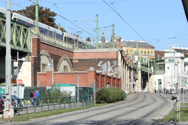 Stadtstraßenszene mit einem Zug auf Schienen, umgeben von Gebäuden, Straßeninfrastruktur, Fahrzeugen, Füßgängern, Vegetation und einem klaren blauen Himmel.