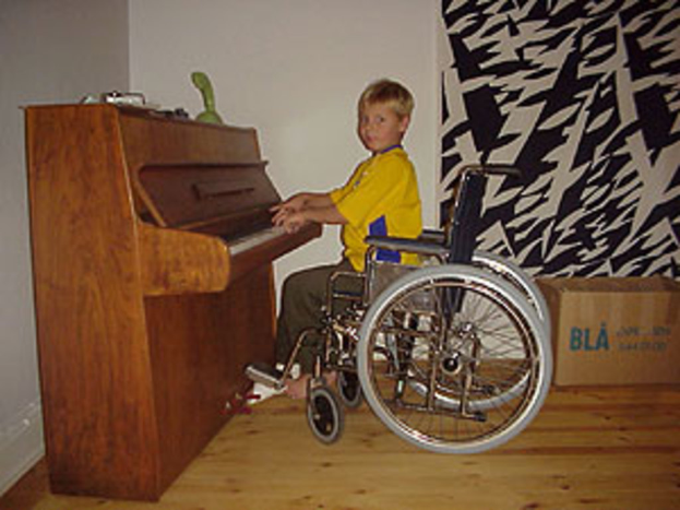 Ein Junge in einem gelben Hemd und Hose sitzt in einem Rollstuhl, spielt auf einem Piano mit Spielzeugen darauf, neben einem Vorhang und einer Kiste auf dem Boden.
